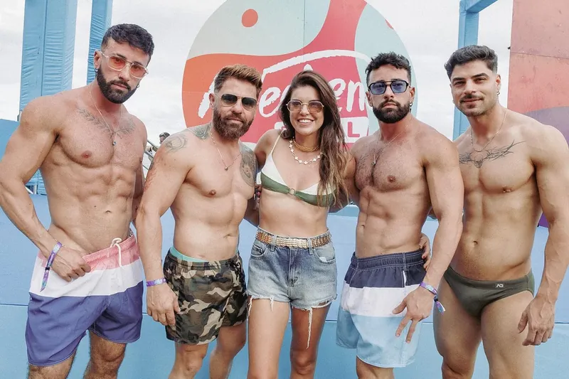Cinco homens sem camisa, de sunga e bermuda, abraçados a uma mulher de biquíni diante do logo do Weekend Festival