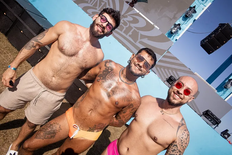 Três homens sem camisa, de sunga e short, sorrindo e posando juntos diante do palco