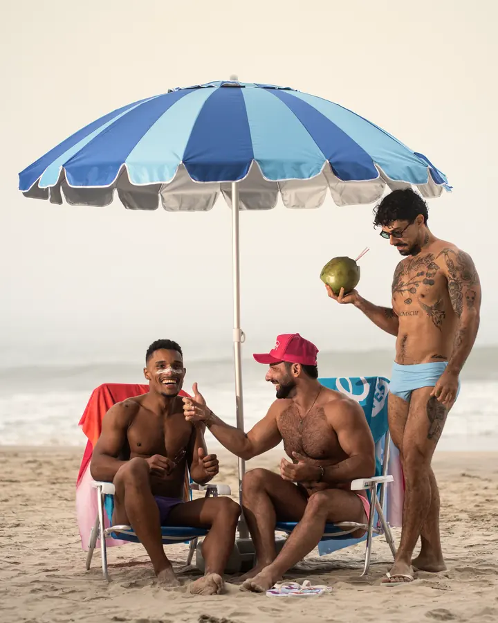 Três amigos de sunga sob o guarda-sol na areia, rindo, um deles com um coco na mão